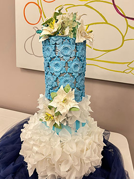 Tall decorative wedding cake with a blue floral-patterned upper tier and voluminous white ruffle-like bottom tier, accented with white and yellow sugar flowers. Displayed on a table with a modern abstract art background.