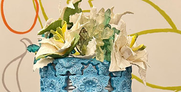 Tall decorative cake with a textured blue upper tier featuring ornate patterns, topped with white and yellow sugar flowers and leaves, set against an abstract background.