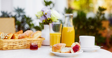 Outdoor breakfast setting with a plate of bread and jam, a basket of pastries, orange juice, and coffee on a white table.