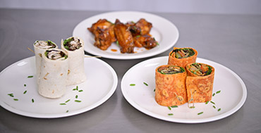 Plates with vegetable tortilla wraps in foreground and glazed chicken wings in background.