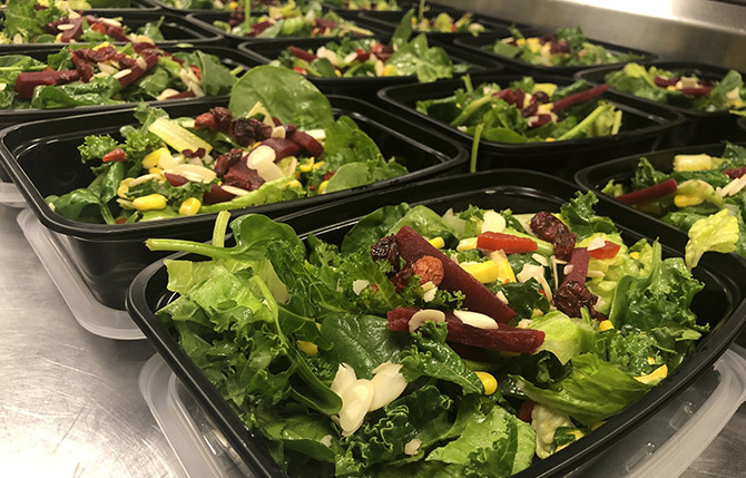 Rows of fresh green salads in black meal prep containers with beets, corn, and sliced almonds.
