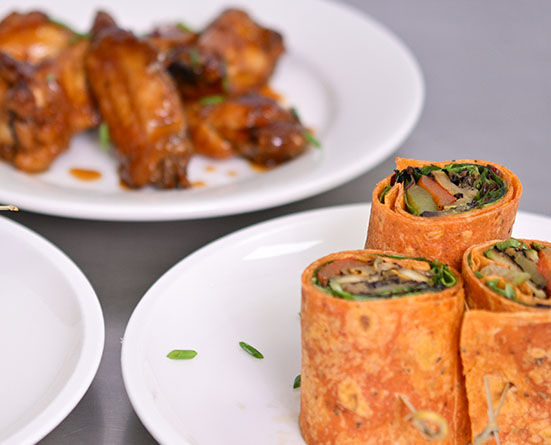 Plates with vegetable tortilla wraps in foreground and glazed chicken wings in background.