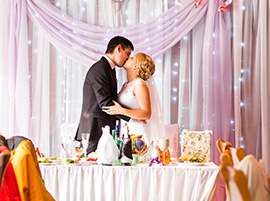 Bride and groom sharing a kiss at their decorated reception table with draped lights and pastel curtains.